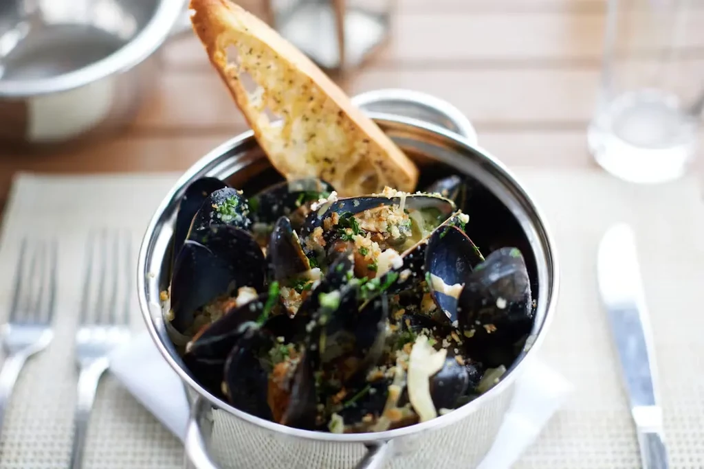 A metal pot of mussels with herbs and a slice of garlic bread.