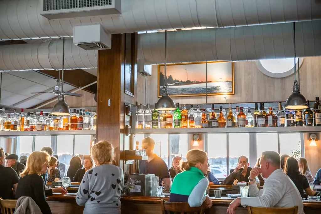 Patrons seated at a bar with many liquor bottles and a sunset painting.