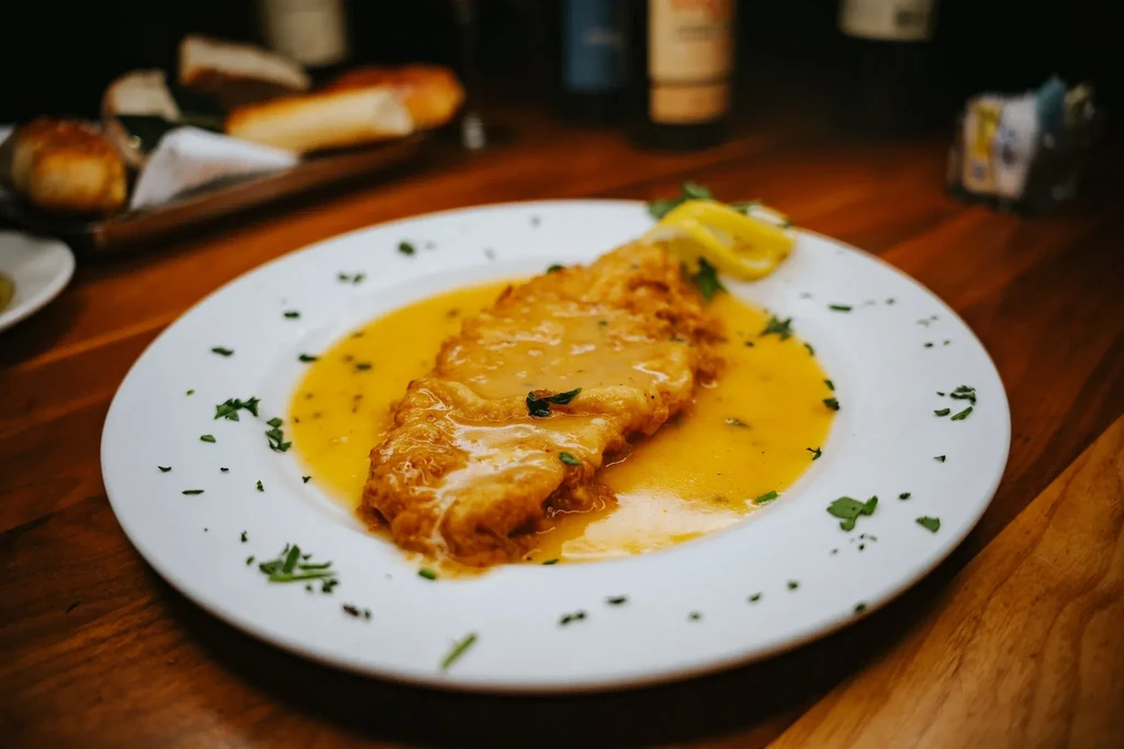 A breaded cutlet in yellow sauce with parsley on a white plate.