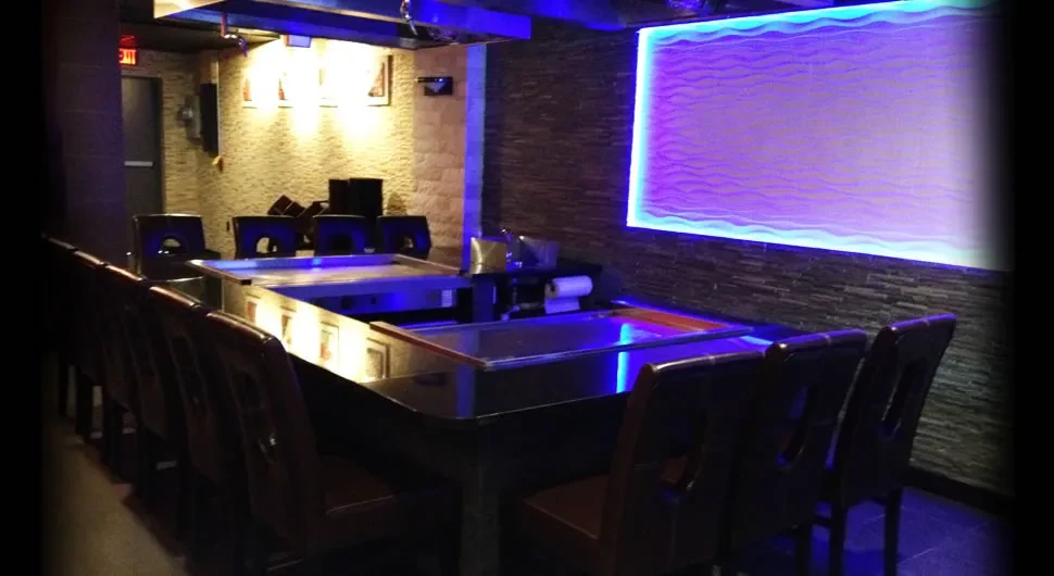 Teppanyaki restaurant with dark chairs around built-in cooking surfaces and purple-lit walls.