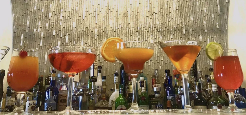 Five colorful cocktails, garnished, on a bar with liquor bottles.