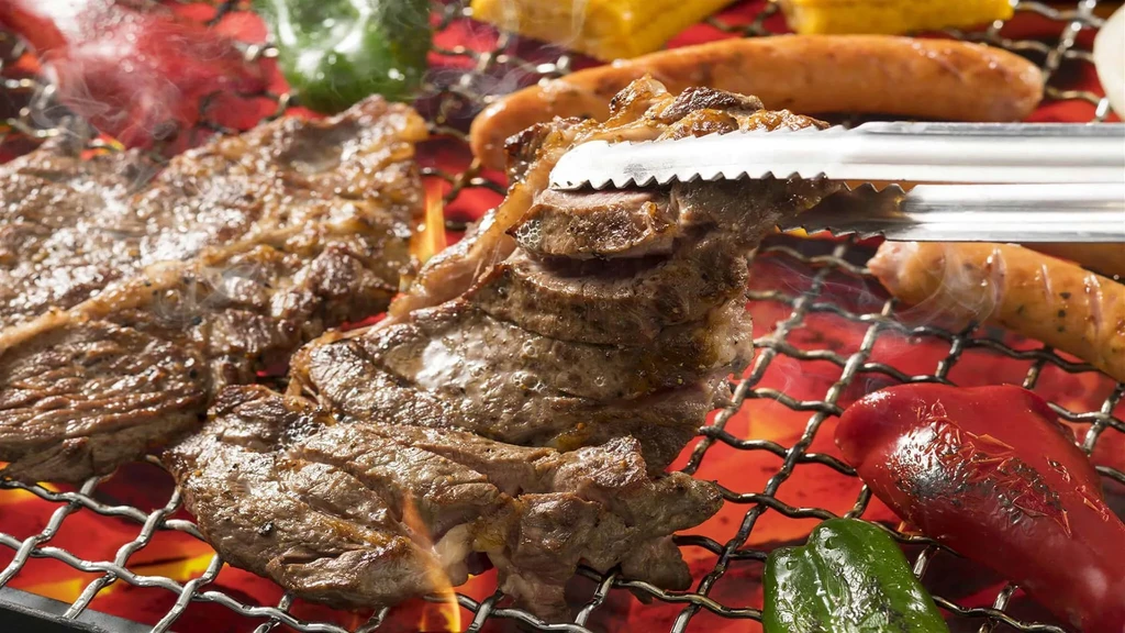 A hand uses tongs to flip meat on a barbecue grill.