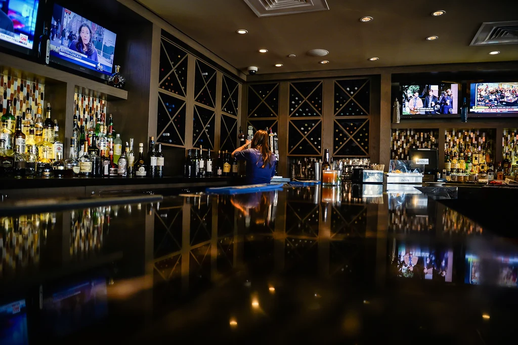 Bartender behind a fully stocked bar with TVs and wine racks.