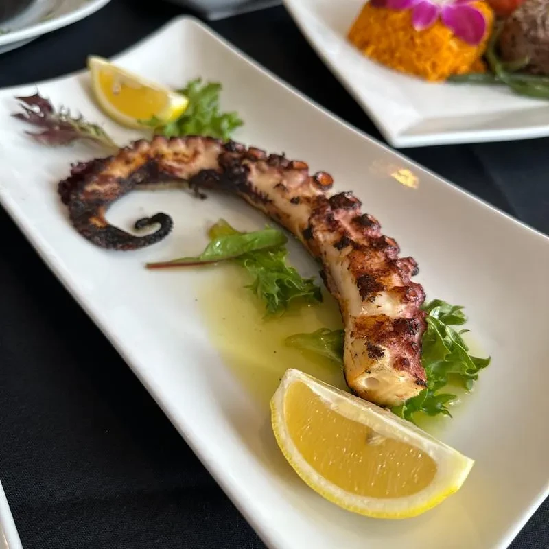 Grilled octopus leg with lemon wedges, lettuce, and oil on white plate.
