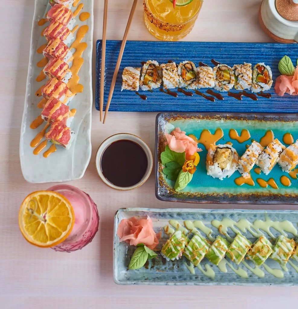 An overhead shot of four colorful sushi platters with drinks and sauce.
