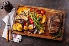 Grilled steak and vegetables served on a wooden board with cutlery.