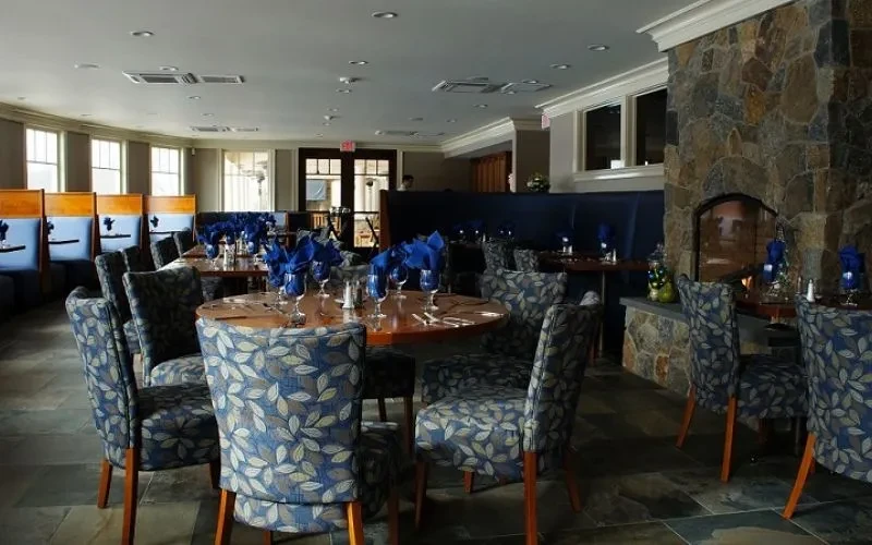 Bright restaurant dining room with tables, blue chairs, booths, stone fireplace.