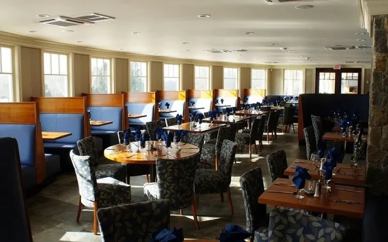 Sunny restaurant dining room with blue booths, set tables, stone wall.