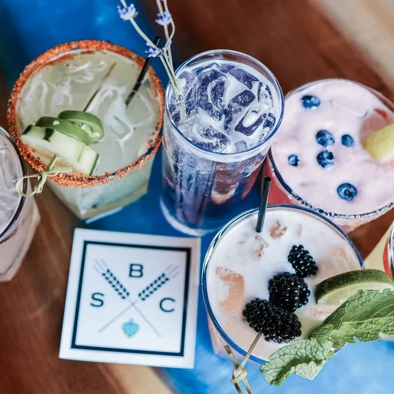 Four colorful cocktails, garnished with fruit, herbs, and spices, on a table.