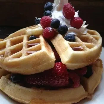 Stacked golden-brown waffles with fresh berries, whipped cream, and syrup drizzle.
