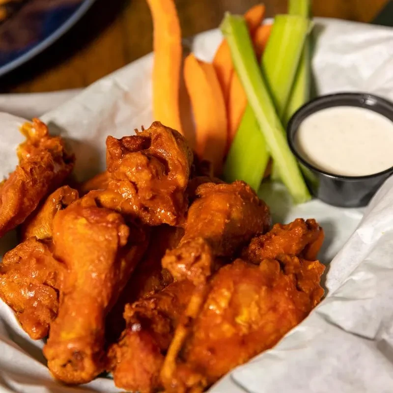 Basket of bright orange buffalo wings with celery, carrots, and dipping sauce.