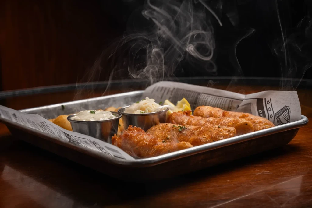 Steaming fish and chips, sauce, coleslaw, lemon on newspaper-lined tray.