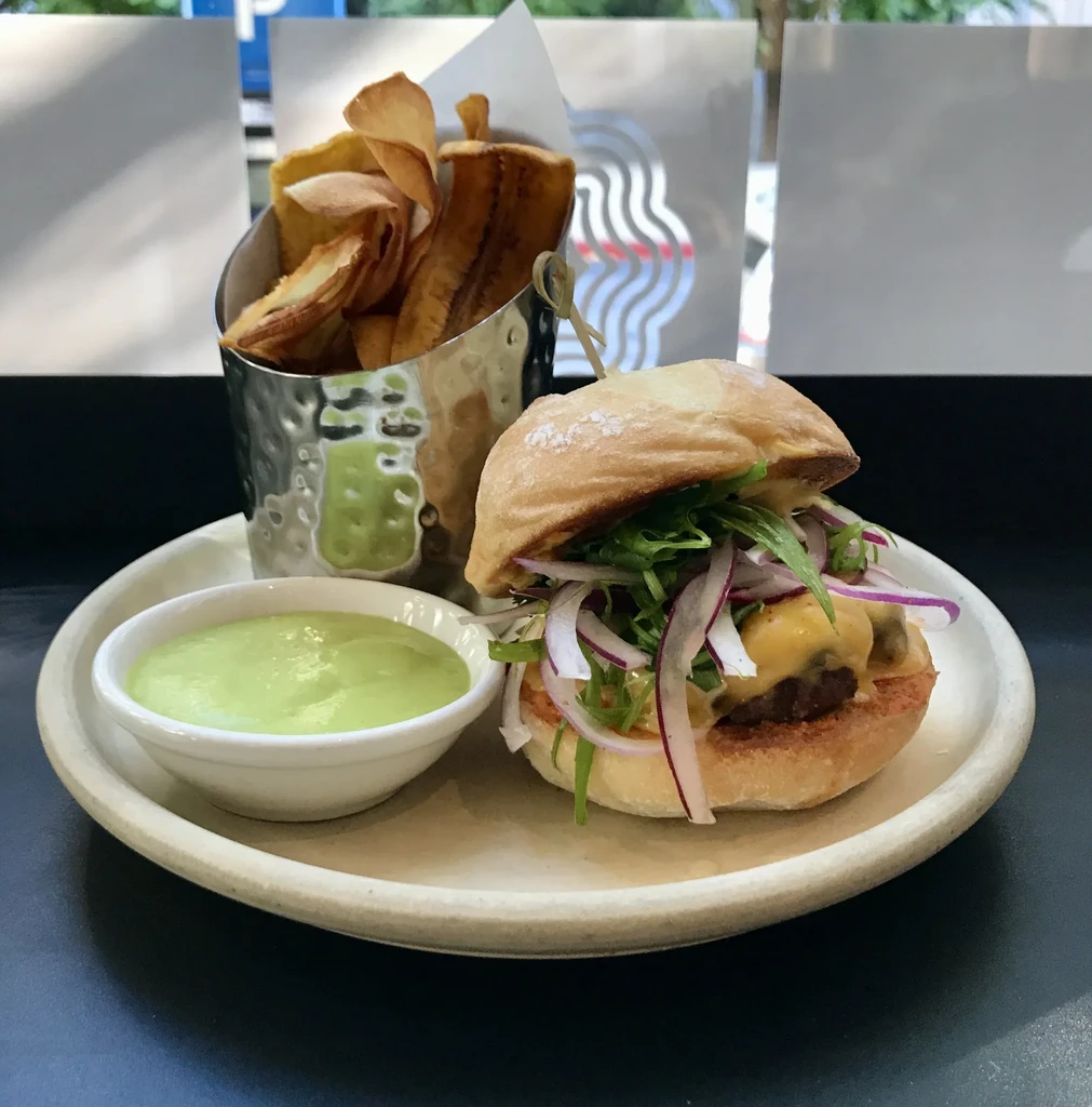 Gourmet burger with cheese and greens, plantain chips, and green dipping sauce.