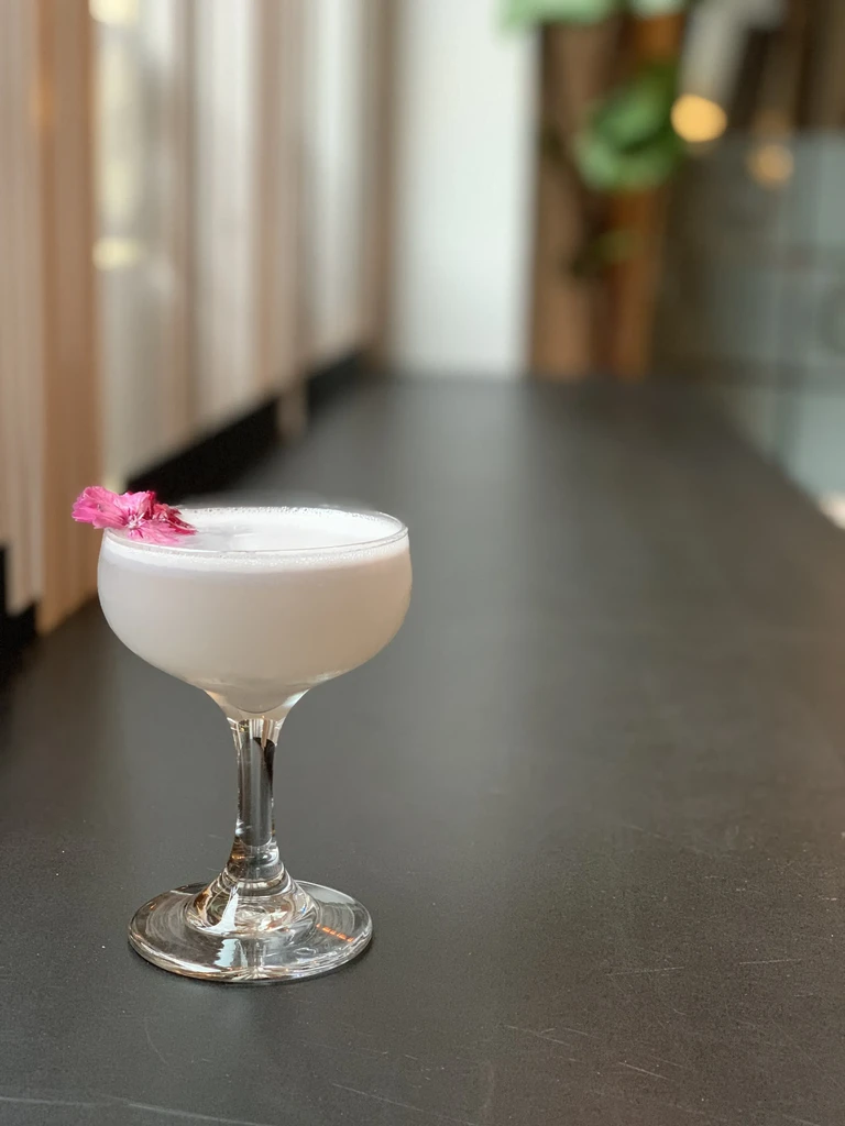 Pale purple cocktail in coupe glass, with foam and pink flower.