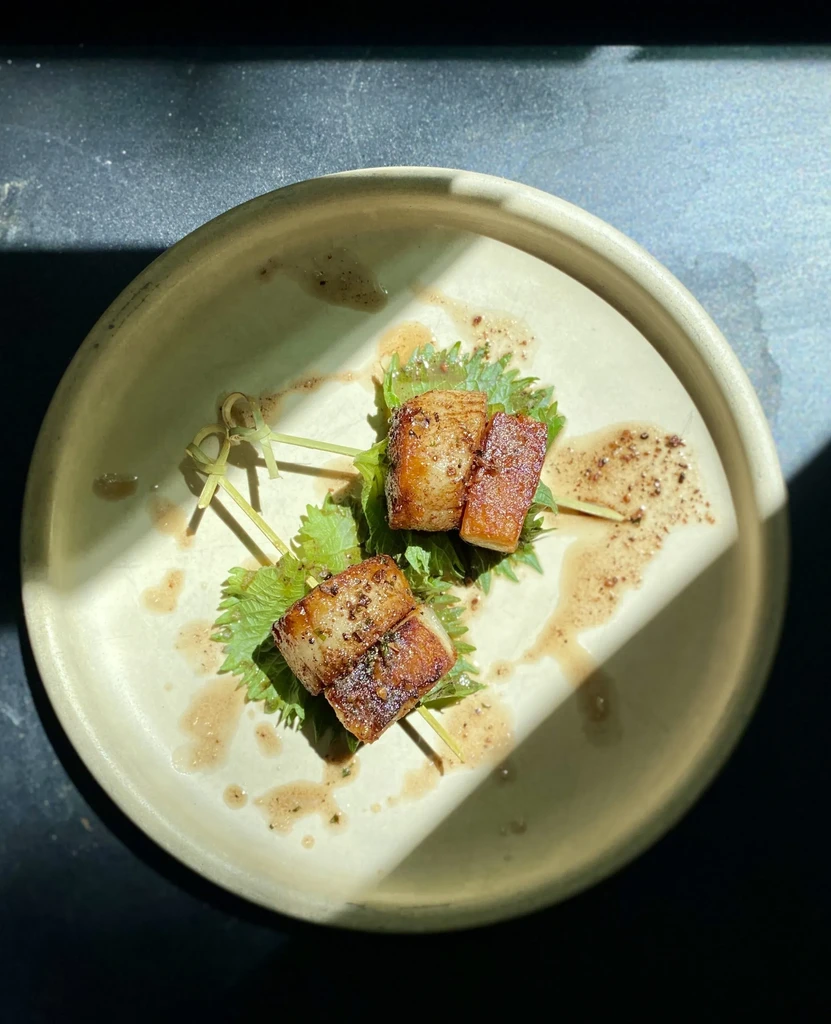Two seared pork belly skewers on a green leaf, drizzled with sauce.