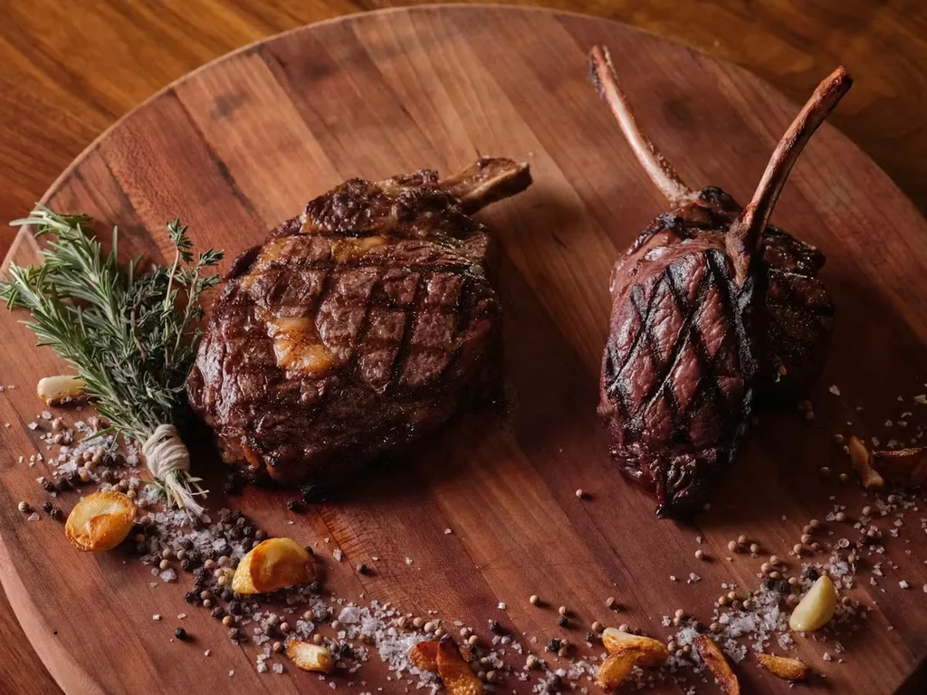 Two grilled bone-in steaks with herbs, garlic, spices on a wooden board.