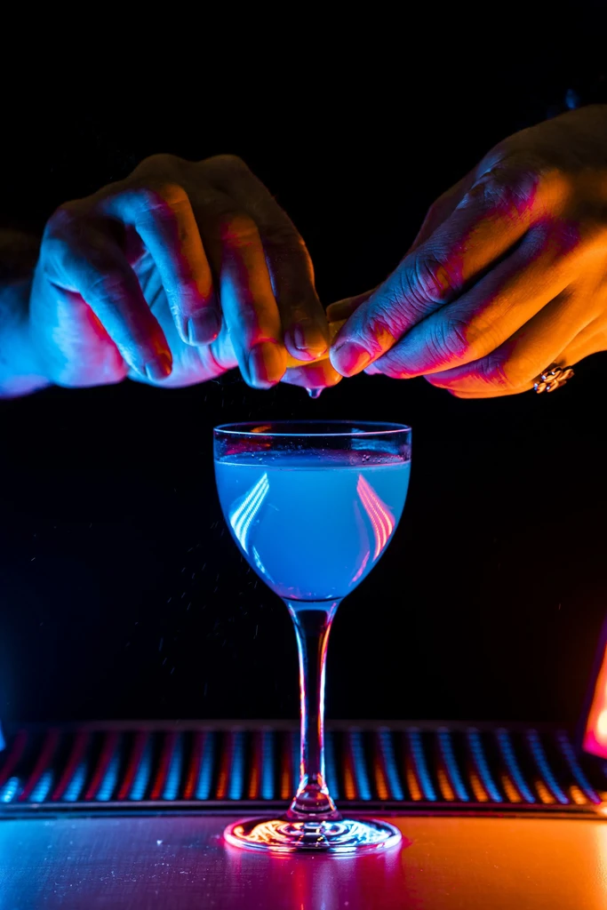 Hands squeeze citrus over blue cocktail under dramatic neon lighting.