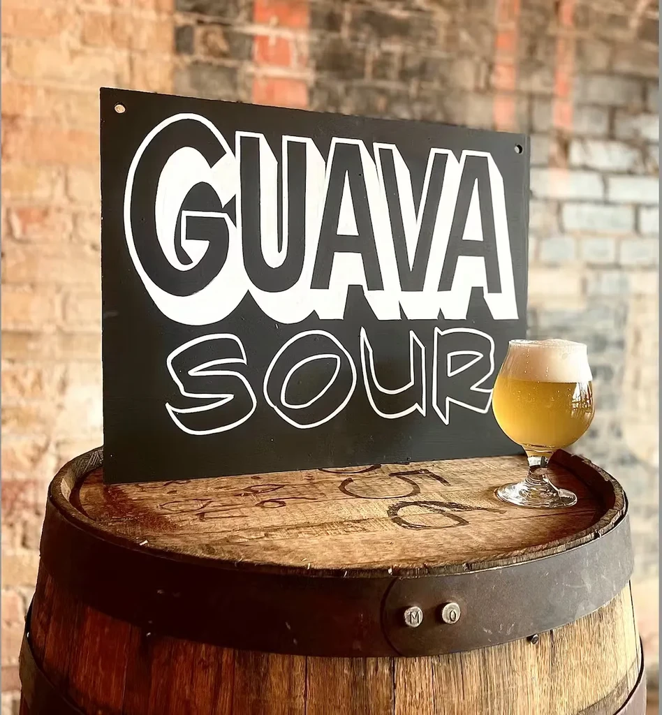 Black "GUAVA SOUR" sign next to a frothy yellow drink on a barrel.