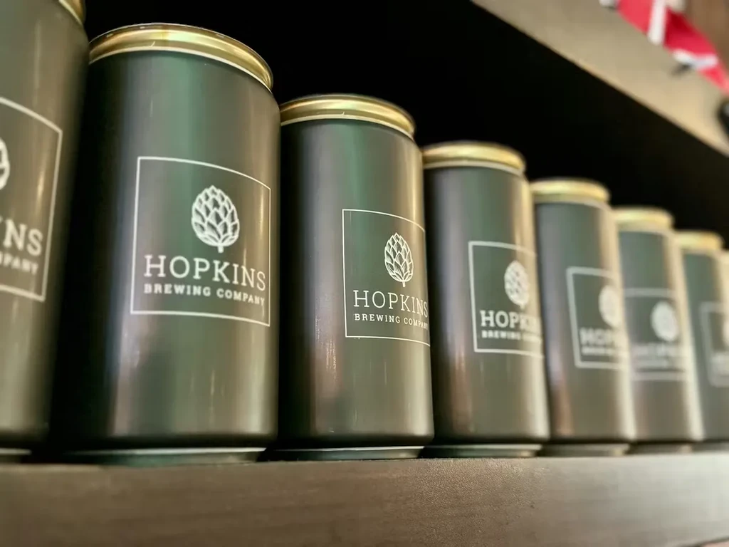 Identical Hopkins Brewing Company cans with gold tops on a dark shelf.