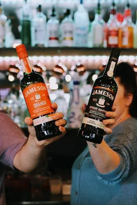 Two hands hold Jameson Orange and Jameson Stout whiskey bottles.