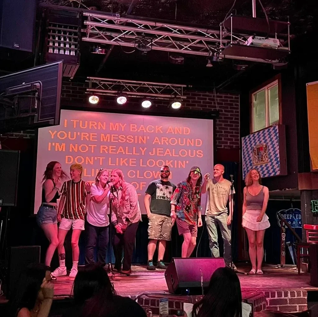 Eight people singing karaoke on stage with pink-purple lights and lyrics.
