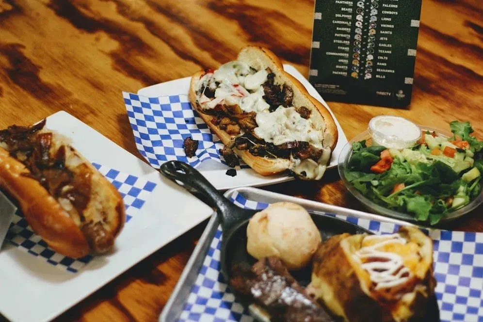 A casual meal of cheesesteak, baked potato, and salad, near sports scores.