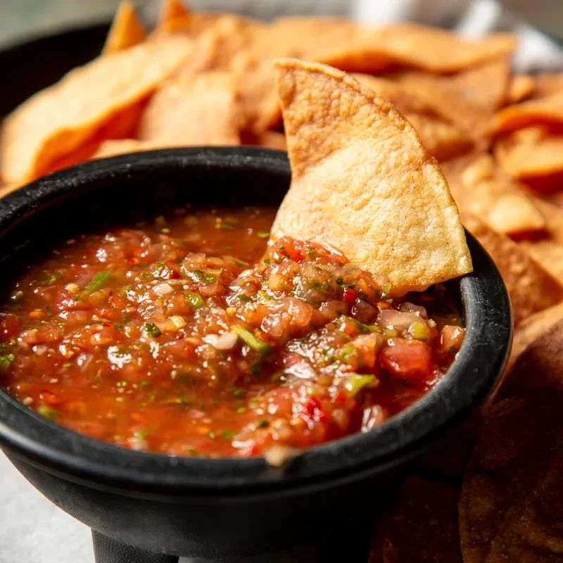 Black bowl of chunky red salsa, a dipping chip, and more chips.