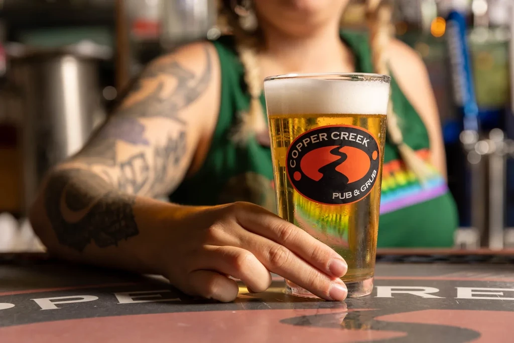 Beer with "Copper Creek Pub & Grub" logo, held by a tattooed hand.