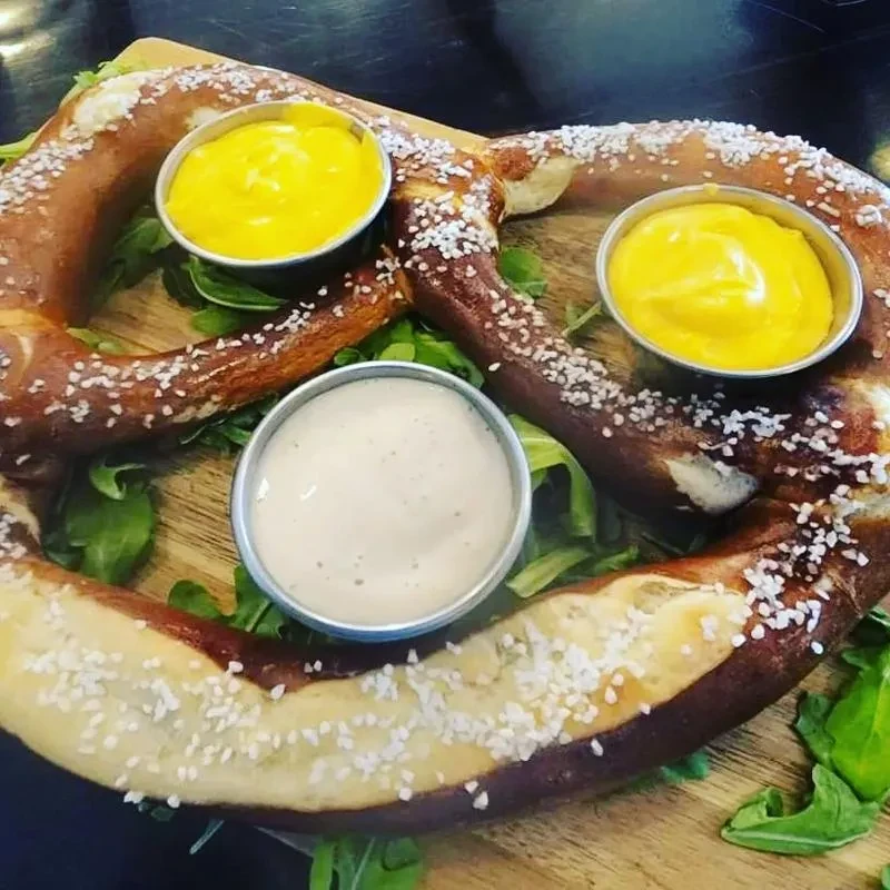 Golden-brown salted pretzel, greens, and three dipping sauces on wood board.