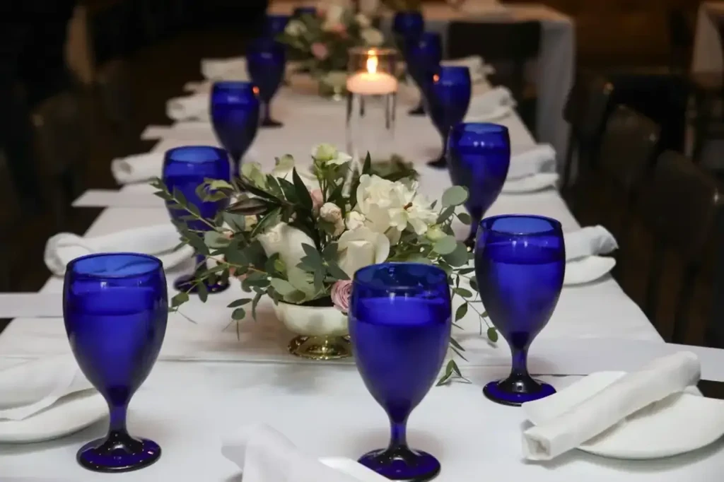 Formal dining table with white settings, blue glasses, and floral centerpiece.