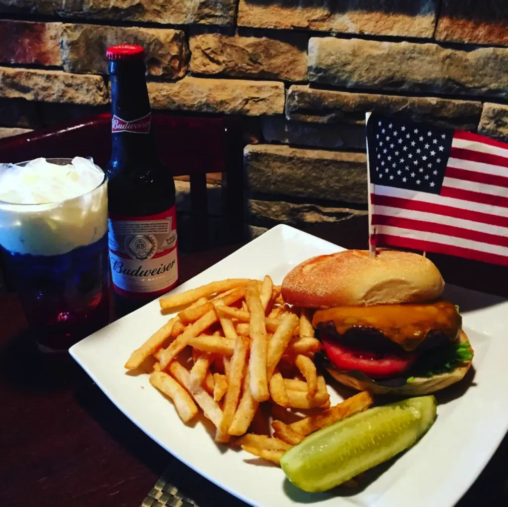 Cheeseburger with American flag, fries, pickle, Budweiser, layered drink.