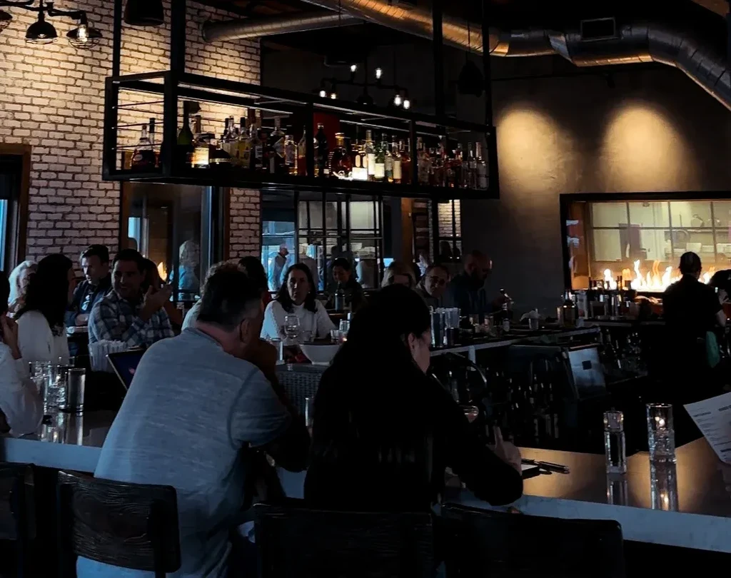 Modern bar interior, bustling with patrons, exposed brick, and open kitchen.
