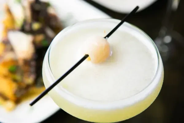 Pale yellow cocktail with frothy white top, garnished with a lychee.