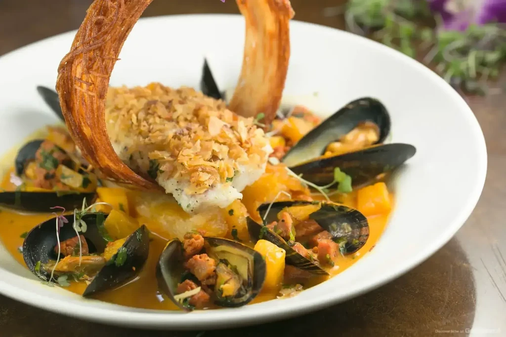 A white bowl of seafood stew with mussels and a crispy fish.