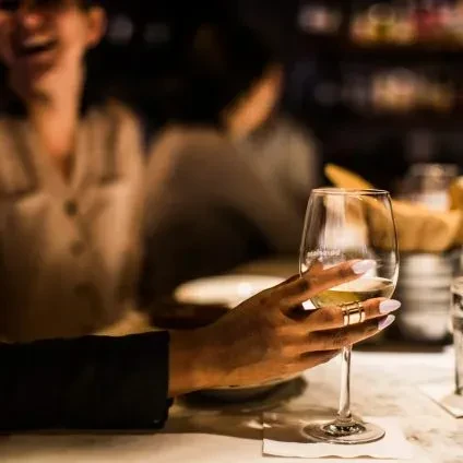 Hand with gold ring holds white wine glass in dimly lit restaurant.