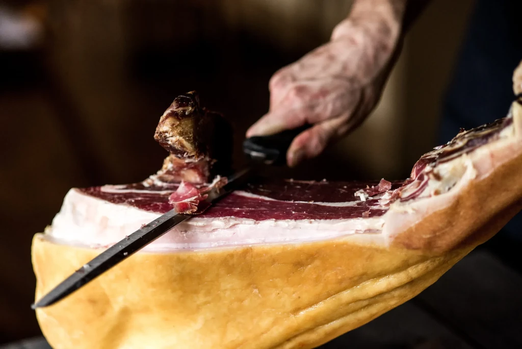 Close-up: Hand slicing cured ham leg, showing red meat and white fat.