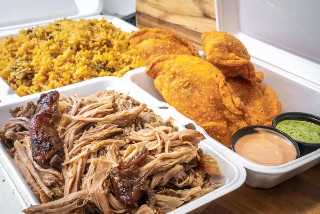 Three takeout containers with rice, pulled pork, empanadas, and dipping sauces.