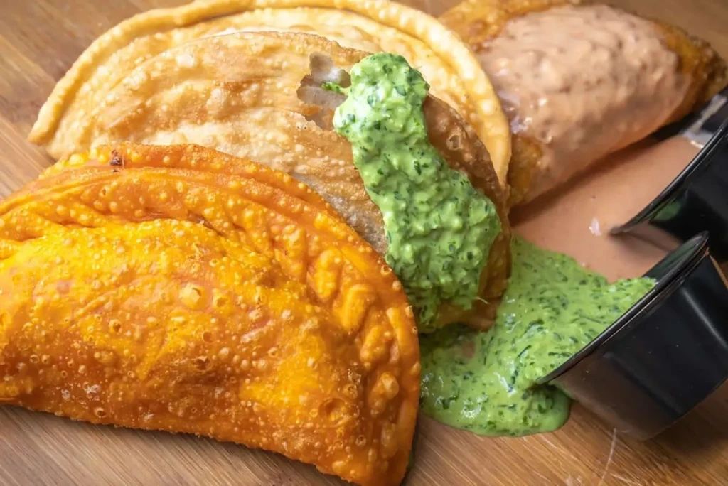 Three fried empanadas with green and red sauces overflowing on a wooden board.