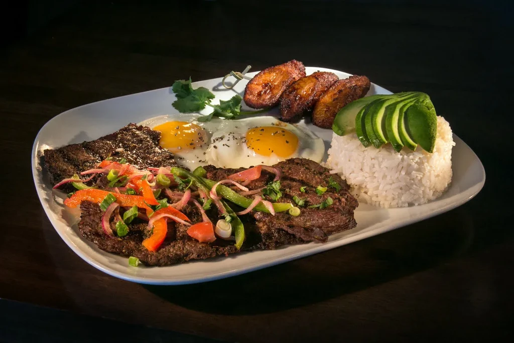 Steak, fried eggs, rice, avocado, and plantains on a white plate.