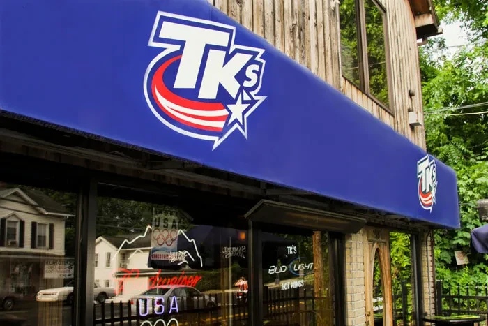 TK's bar exterior: blue awning, wooden siding, neon signs, reflections.