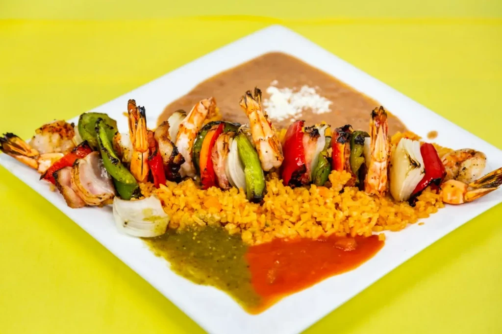 Grilled shrimp and vegetable skewers with orange rice, beans, and sauces.