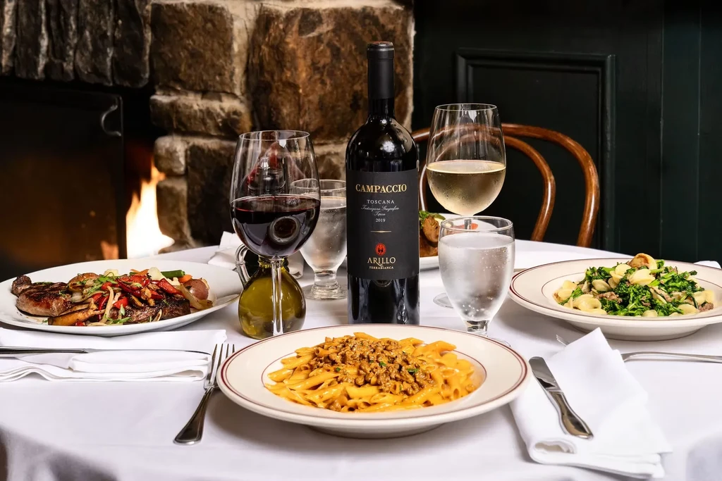Table set with steak, pasta, wine, and water by a fireplace.