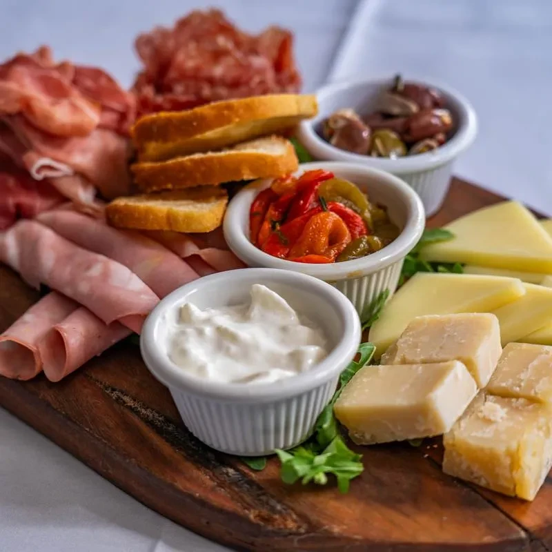 A wooden board with an antipasto platter featuring meats, cheeses, and dips.