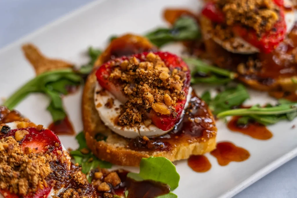 Toasted bread slices topped with strawberries, cheese, arugula, and nuts.
