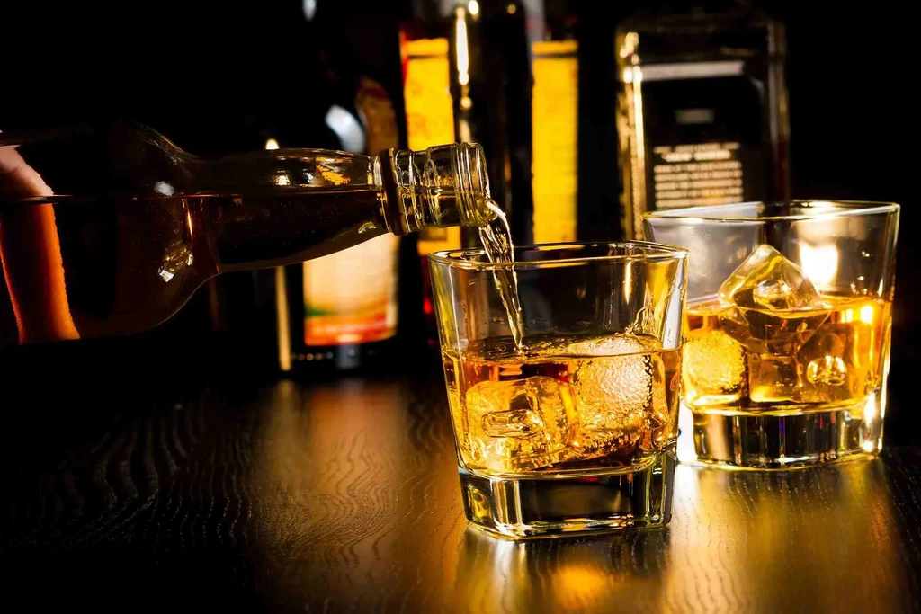 Hand pouring whiskey into an ice-filled glass, second glass nearby.