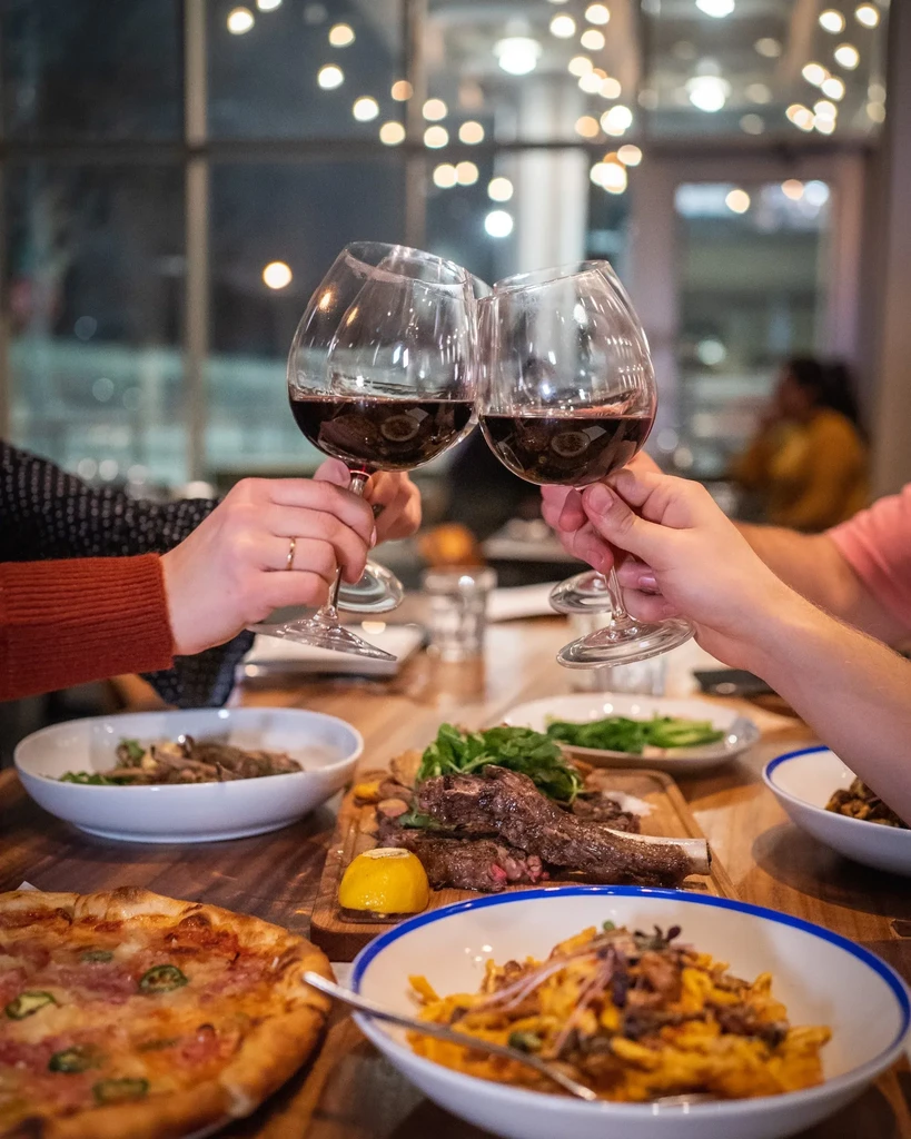 Hands clinking red wine glasses over a table of varied food.