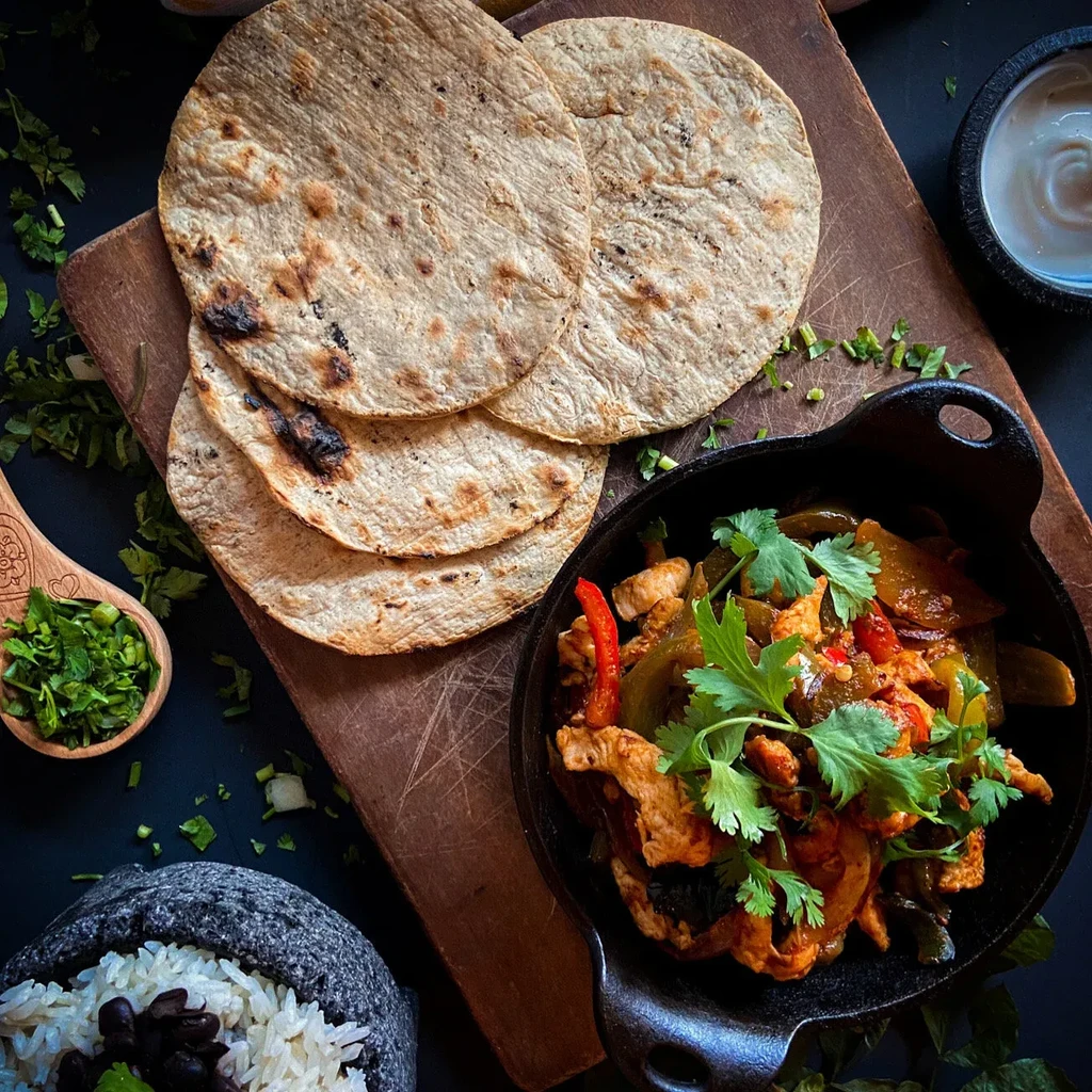 Chicken fajitas with tortillas, rice, and beans on a dark surface.