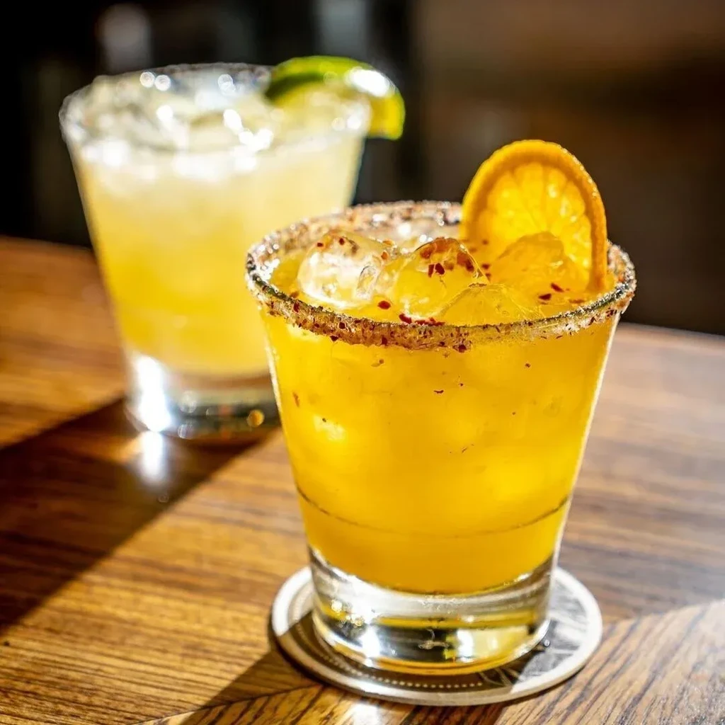 Two cocktails, one with an orange slice and chili flakes, on a wooden table.
