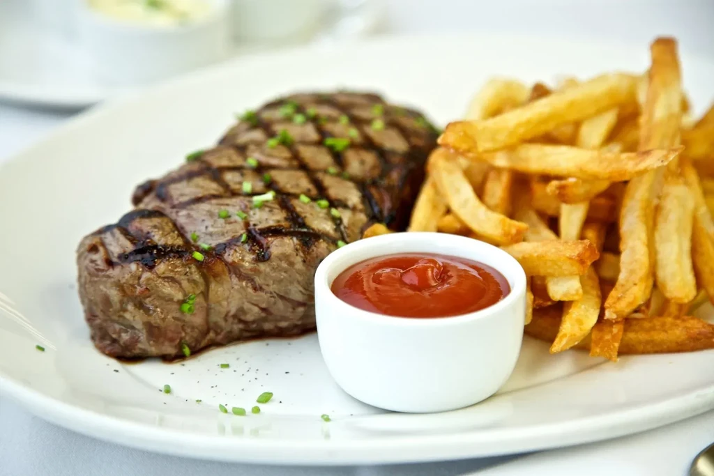 Grilled steak with fries and a small dish of ketchup on a white plate.