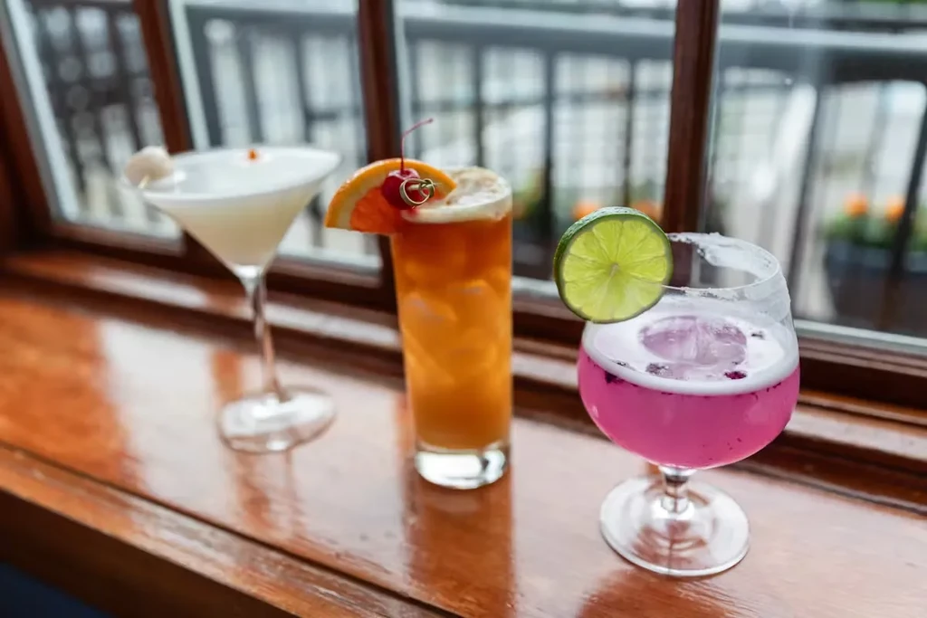 Three colorful cocktails with fruit garnishes sit on a wooden ledge.